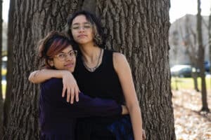 Two teens cuddle and lean into a tree behind them