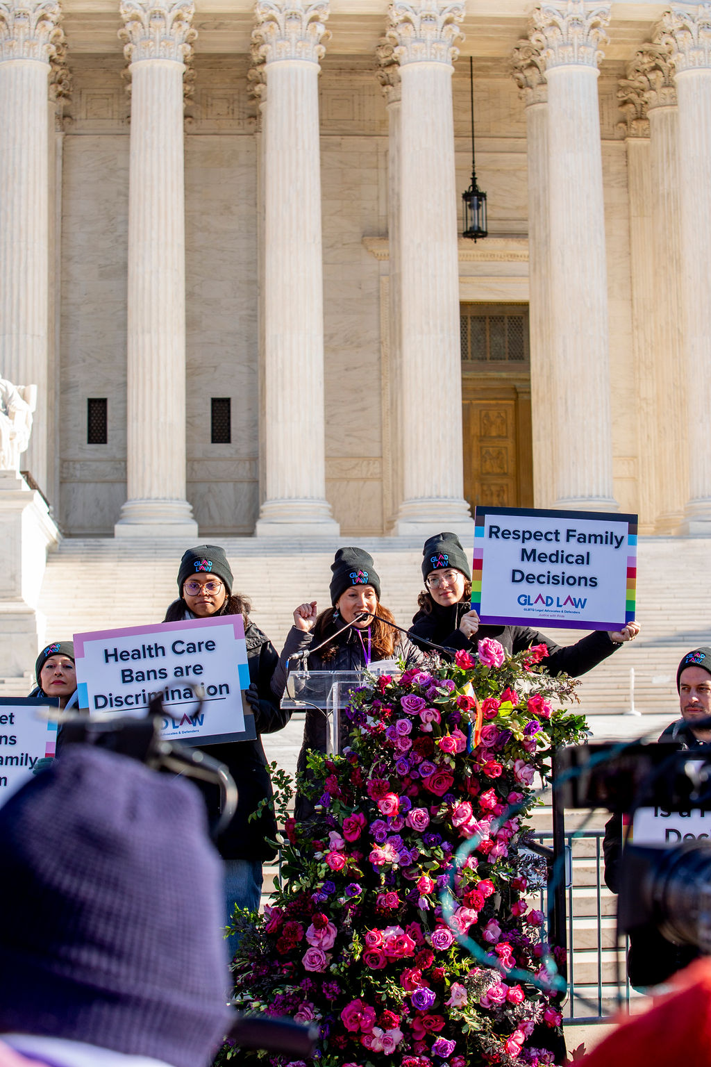 GLAD Law attorney Lisa Rodriguez-Ross speaking at the podium in front of the Supreme Court, surrounded by GLAD Law staff holding signs supporting health care for transgender adolescents.