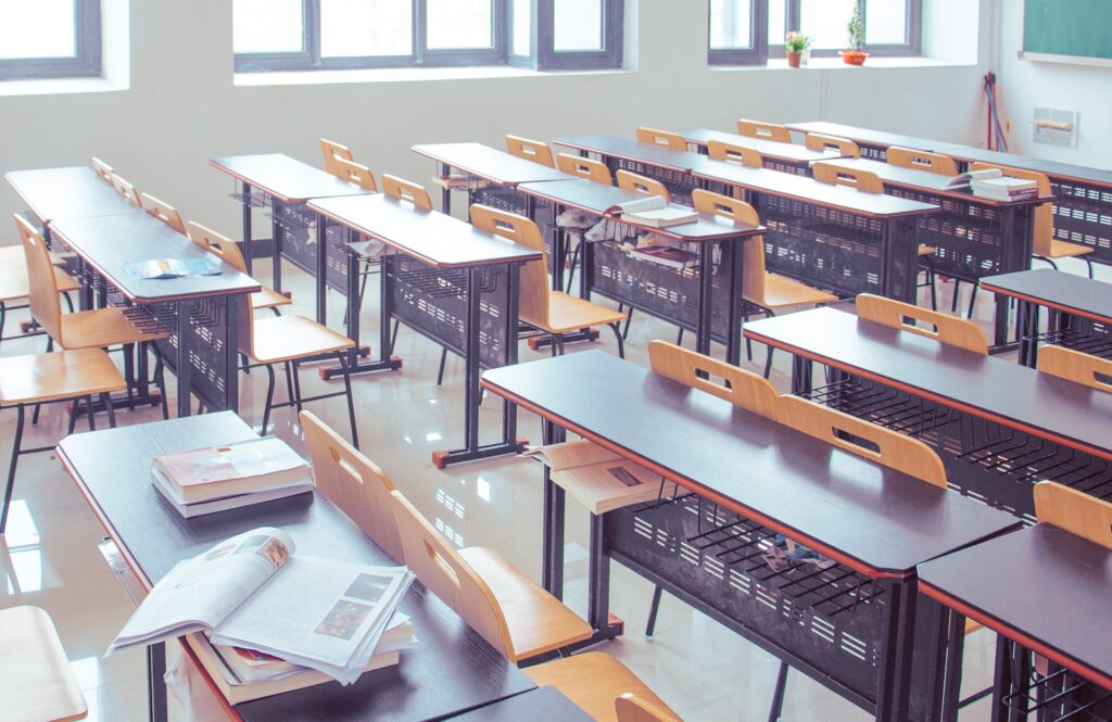 rows of brown table style desks with yellow chairs in a classroom. Some of the desks feature open books.