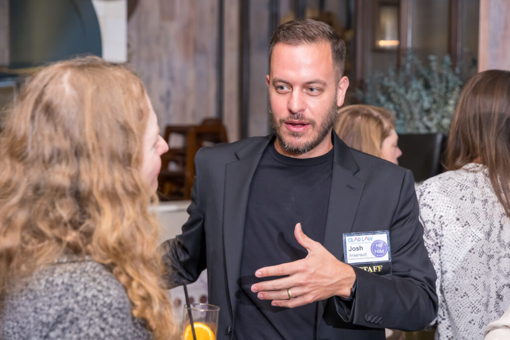 Josh Arsenault at a donor event in a black shirt and blazer, speaking to a woman who is facing away from the camera