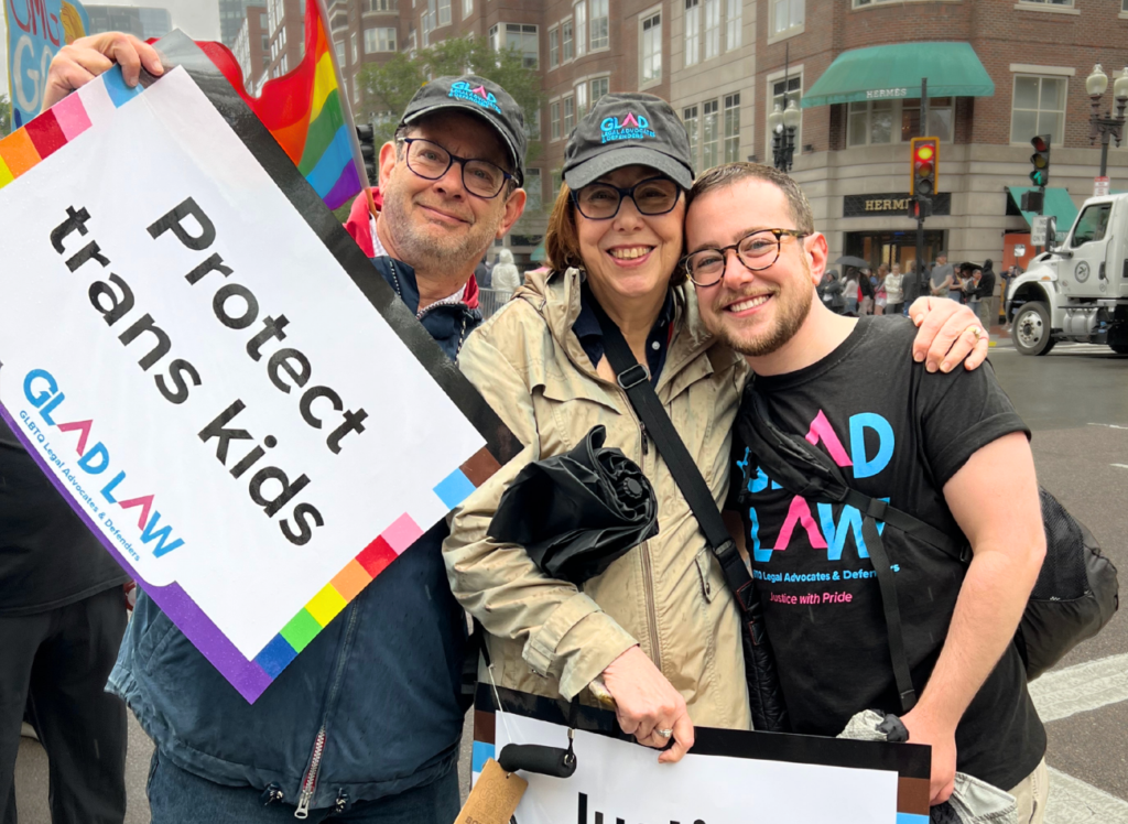Two parents and their adult son are smiling at Boston Pride for the People, holding GLAD Law signs and wearing GLAD Law t-shirts and hats.
