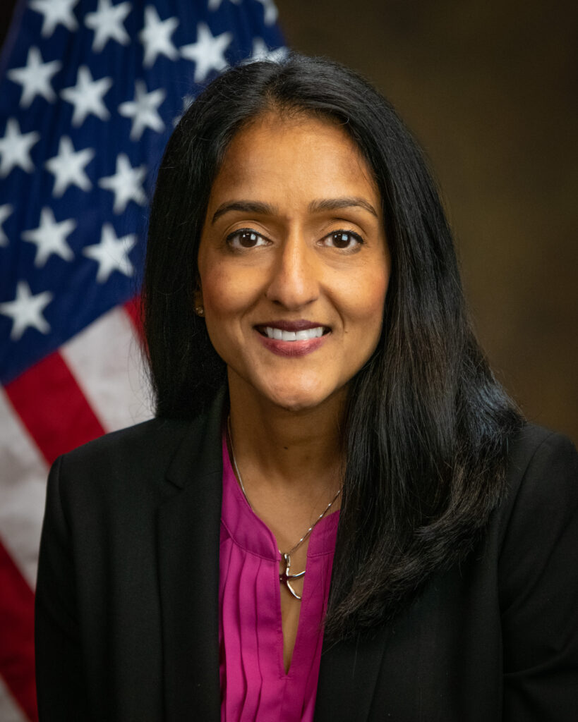 Vanita smiling into the camera, wearing a black blazer over the magenta top, with an American flag in the background