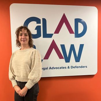 Sky is wearing a white sweater. She is standing in front of a GLAD Law sign