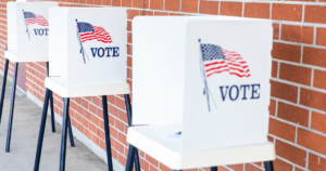 Three white voting booths against a brick wall. Each booth has an American flag and "Vote" on the side.